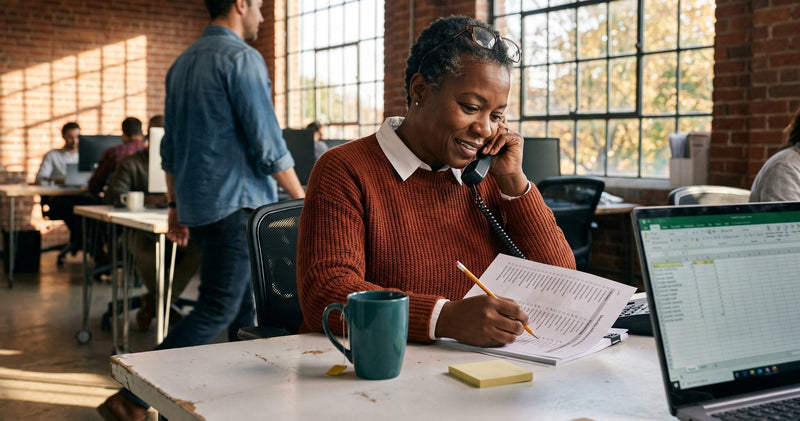 An office manager on a phone call ticks names off a printed recipient list at her desk in an open office.